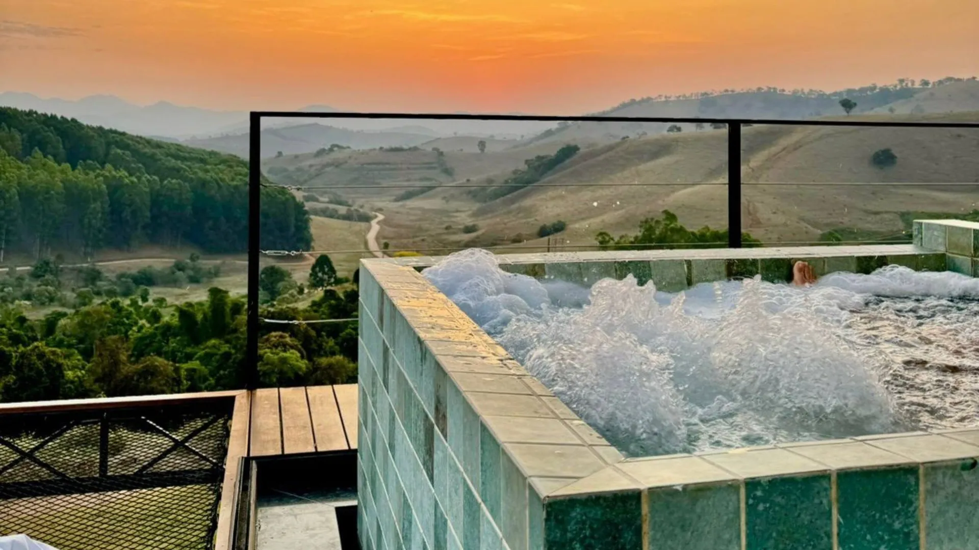 Piscina de borda infinita de azulejo esmeralda numa casa de vidro do Solarium Mantiqueira, com o sol se pondo atrás dos morros da Serra da Mantiqueira